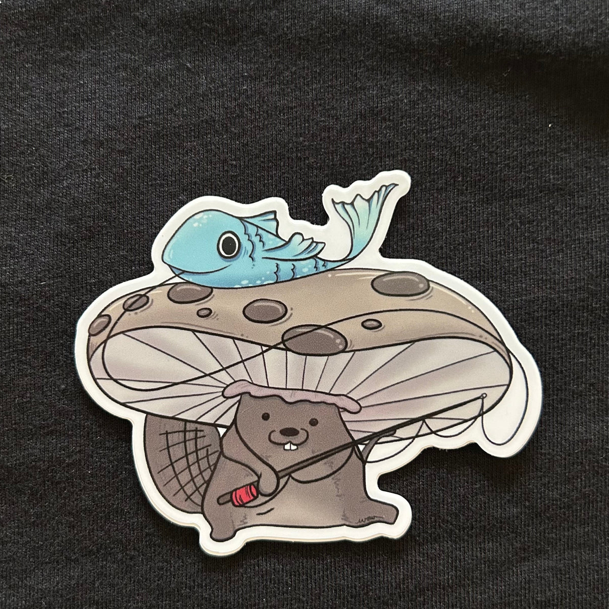 sticker with a beaver wearing a mushroom as a hat and holding a fishing pole. there is a flopping blue fish on top of the mushroom hat. 