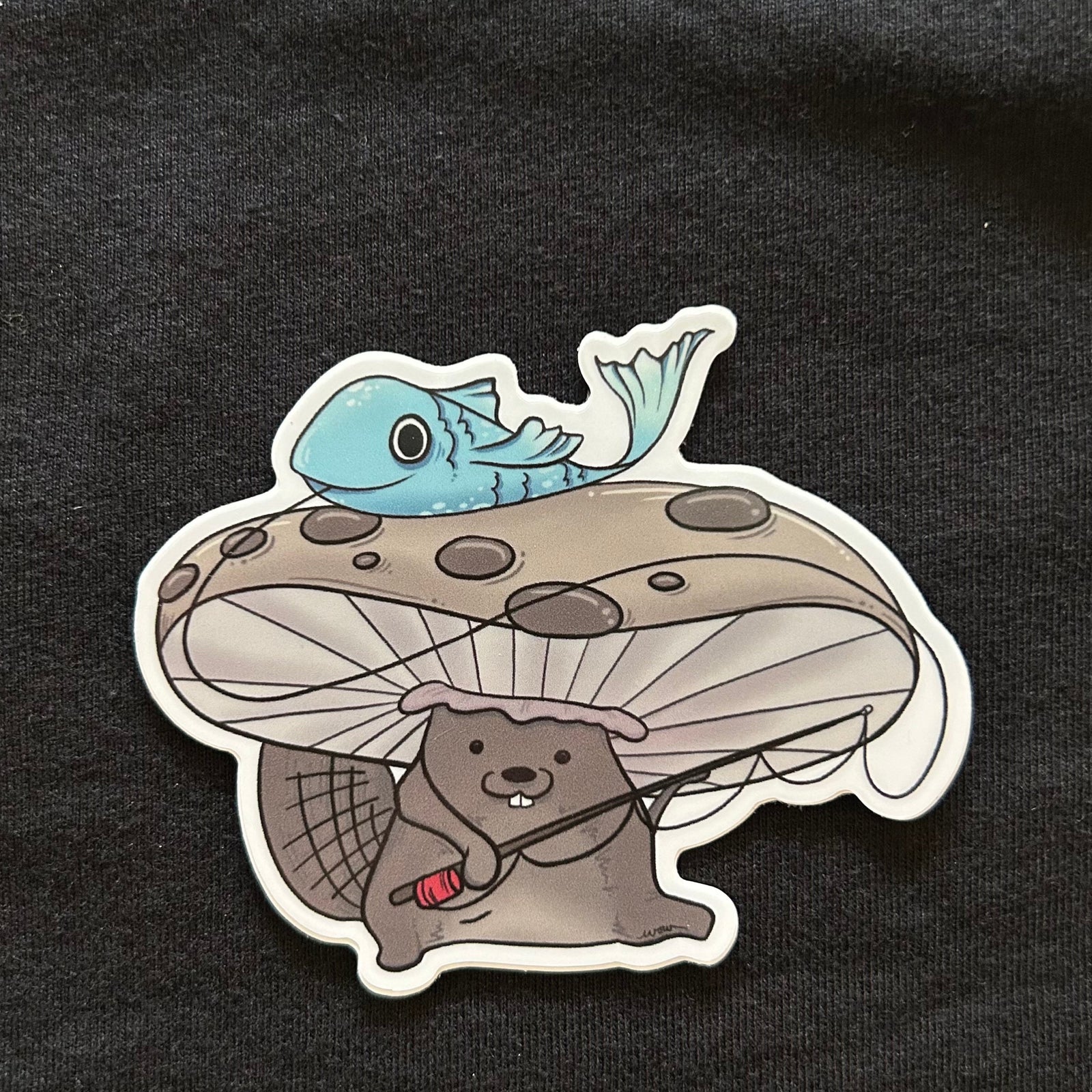 sticker with a beaver wearing a mushroom as a hat and holding a fishing pole. there is a flopping blue fish on top of the mushroom hat. 