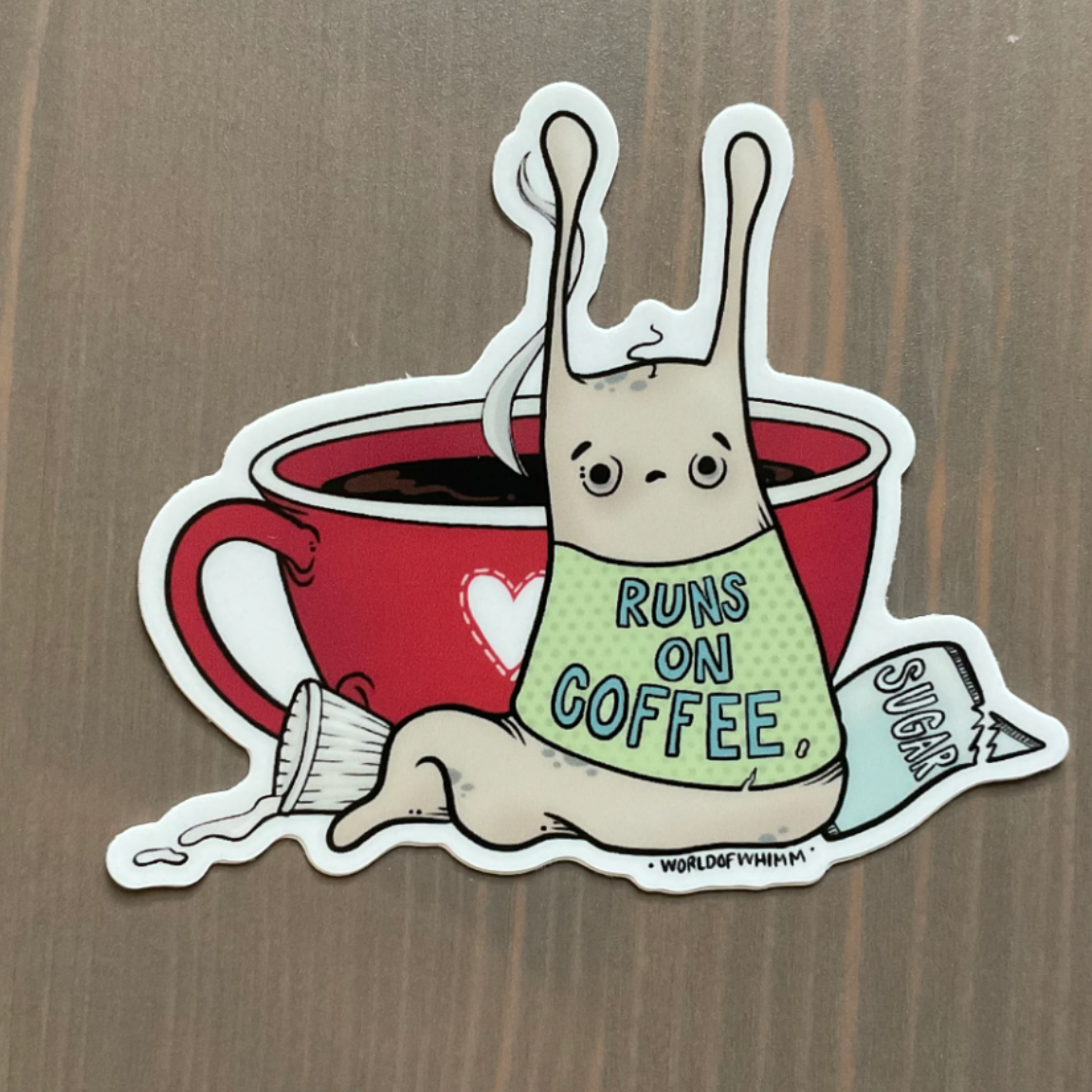 A sticker of a slug wearing a shirt that says "Runs on Coffee" sitting in front of a cup of coffee with a sugar packet and a cup of cream. 