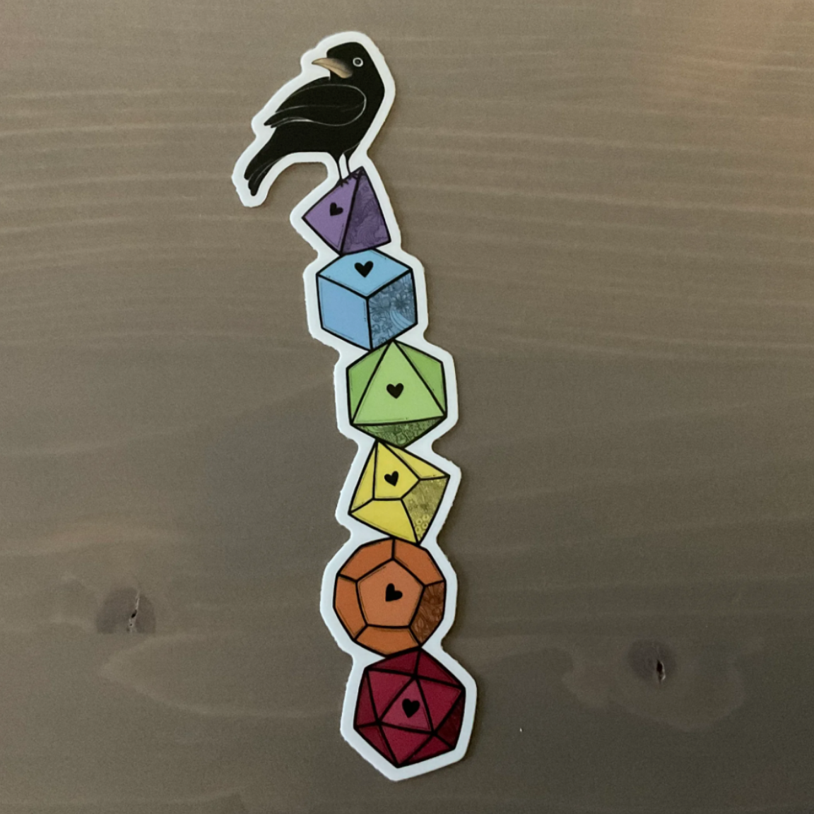 A sticker of a crow sitting on top of a stack of rainbow dice with different numbers of sides.