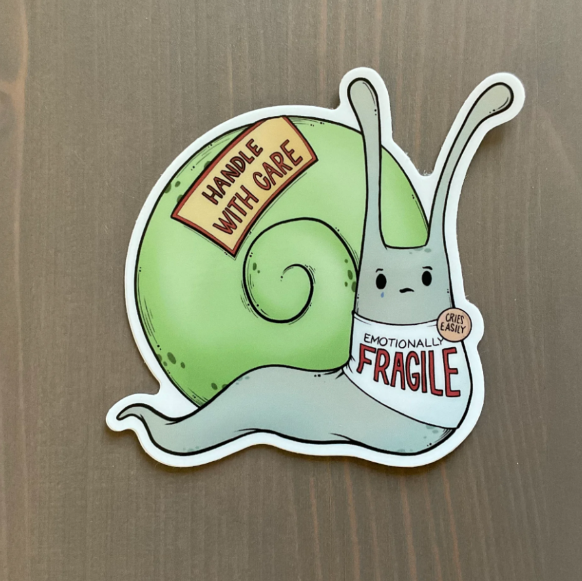 Sticker of a snail with a green shell that is wearing a shirt that says "Emotionally Fragile," a badge that says "Cries Easily," and a sticker on its shell that says "Handle With Care"