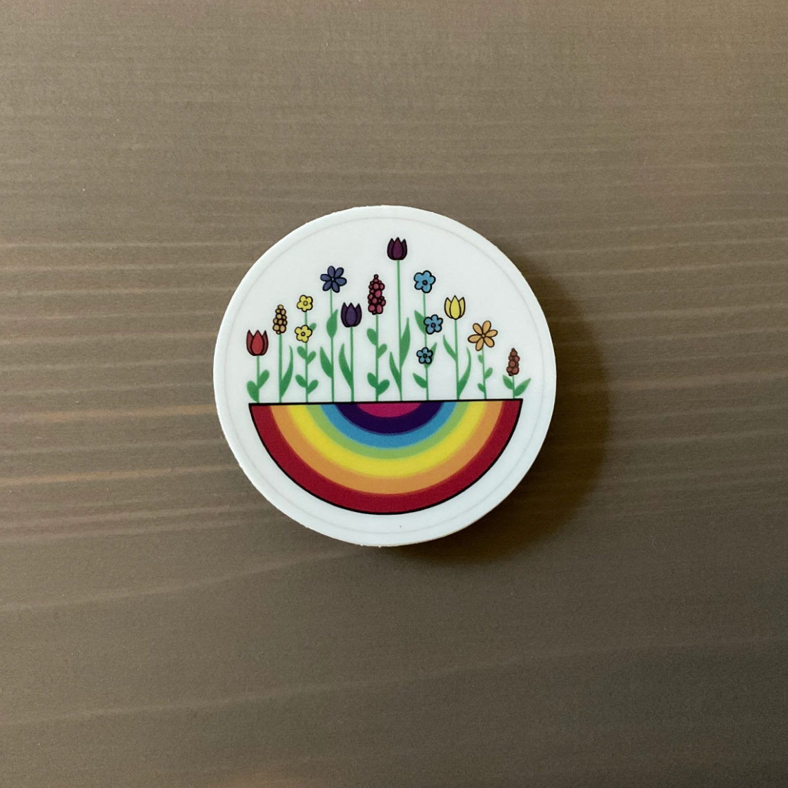A round sticker of a rainbow with multicolor flowers growing out of it