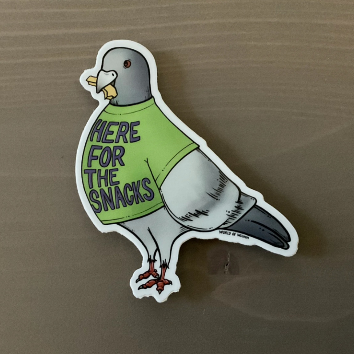 Sticker of a pigeon wearing a green shirt that says "Here for the snacks" on a brown background