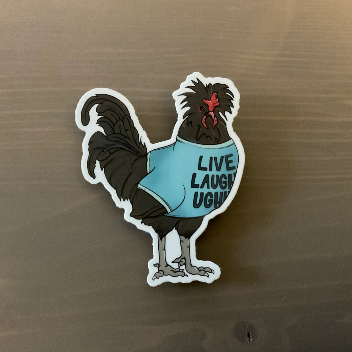 A sticker of a black chicken wearing a blue shirt that says "LIVE LAUGH UGH" on it 