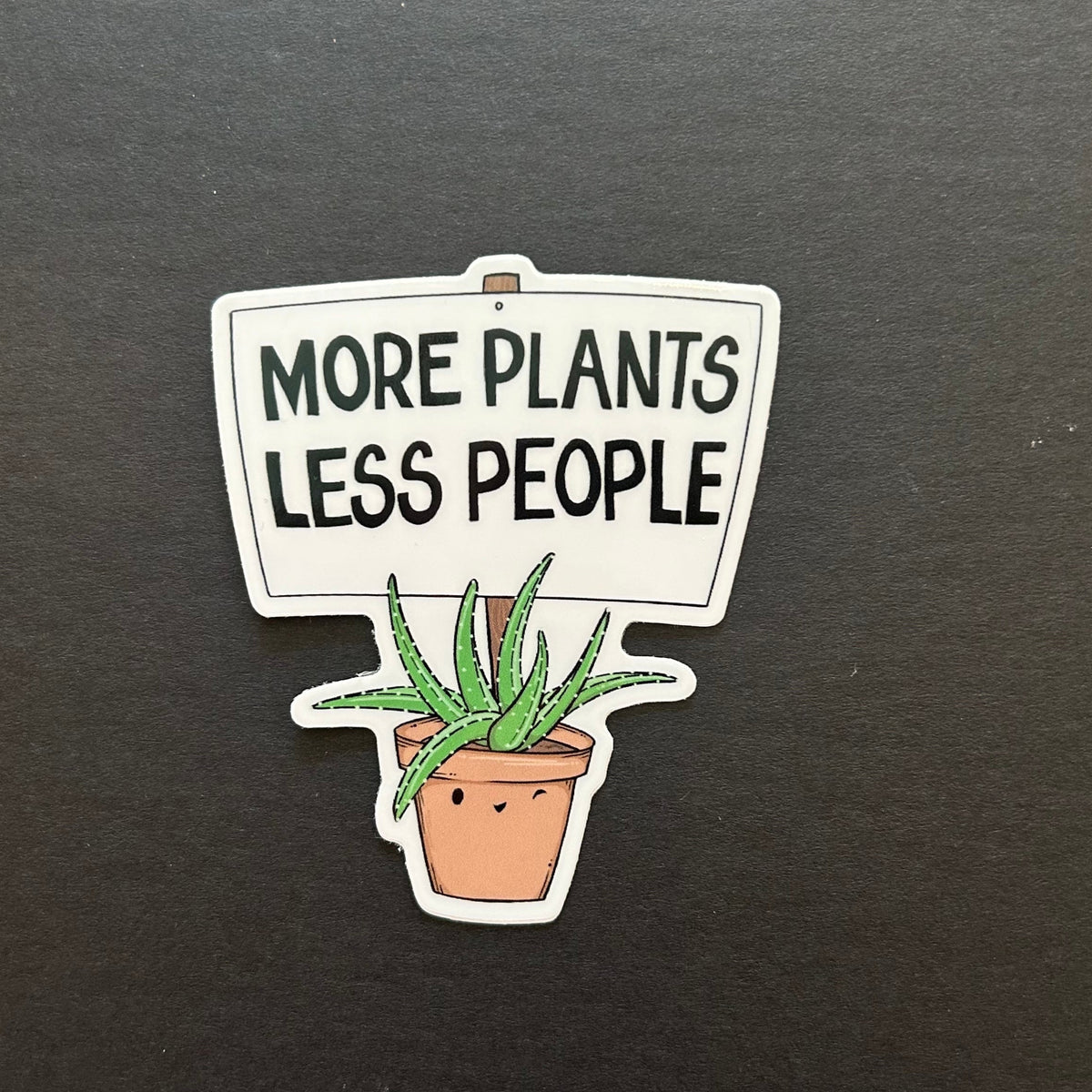 A sticker with a winking aloe plant holding a sign that says "more plants less people"