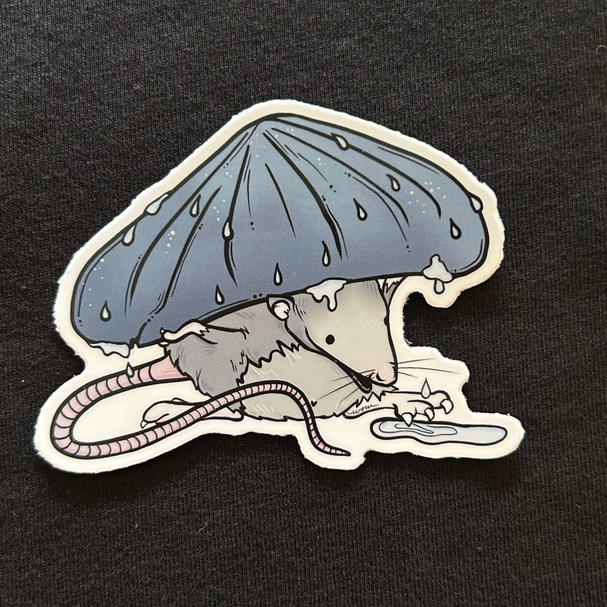 A sticker depicting a possum wearing a mushroom as a hat and splashing its toes in a puddle