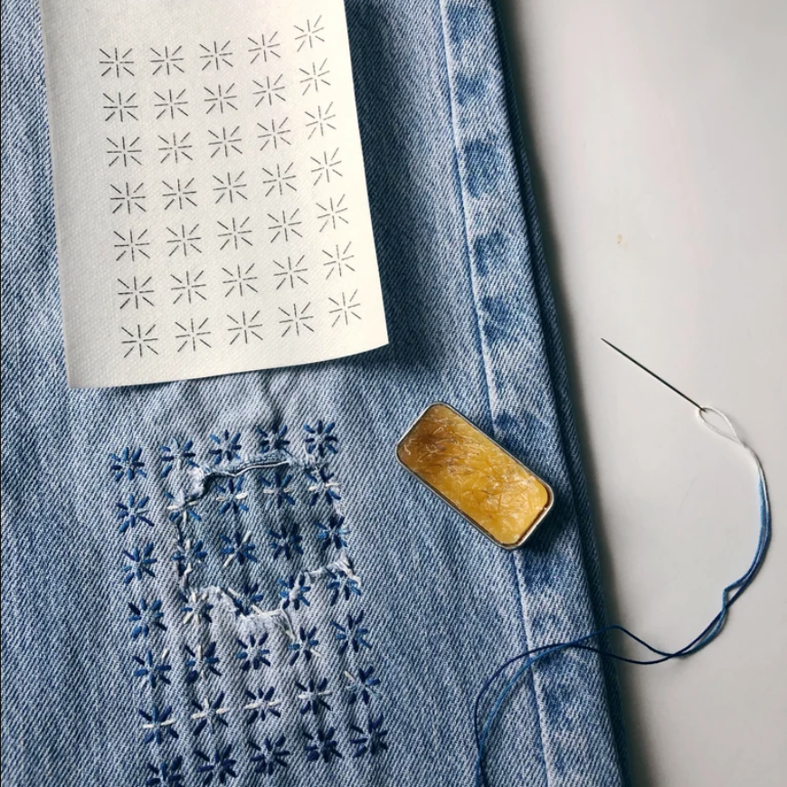 Stitched up example of mending transfers on a pair of jeans with a needle, thread, and tin of thread gloss. 