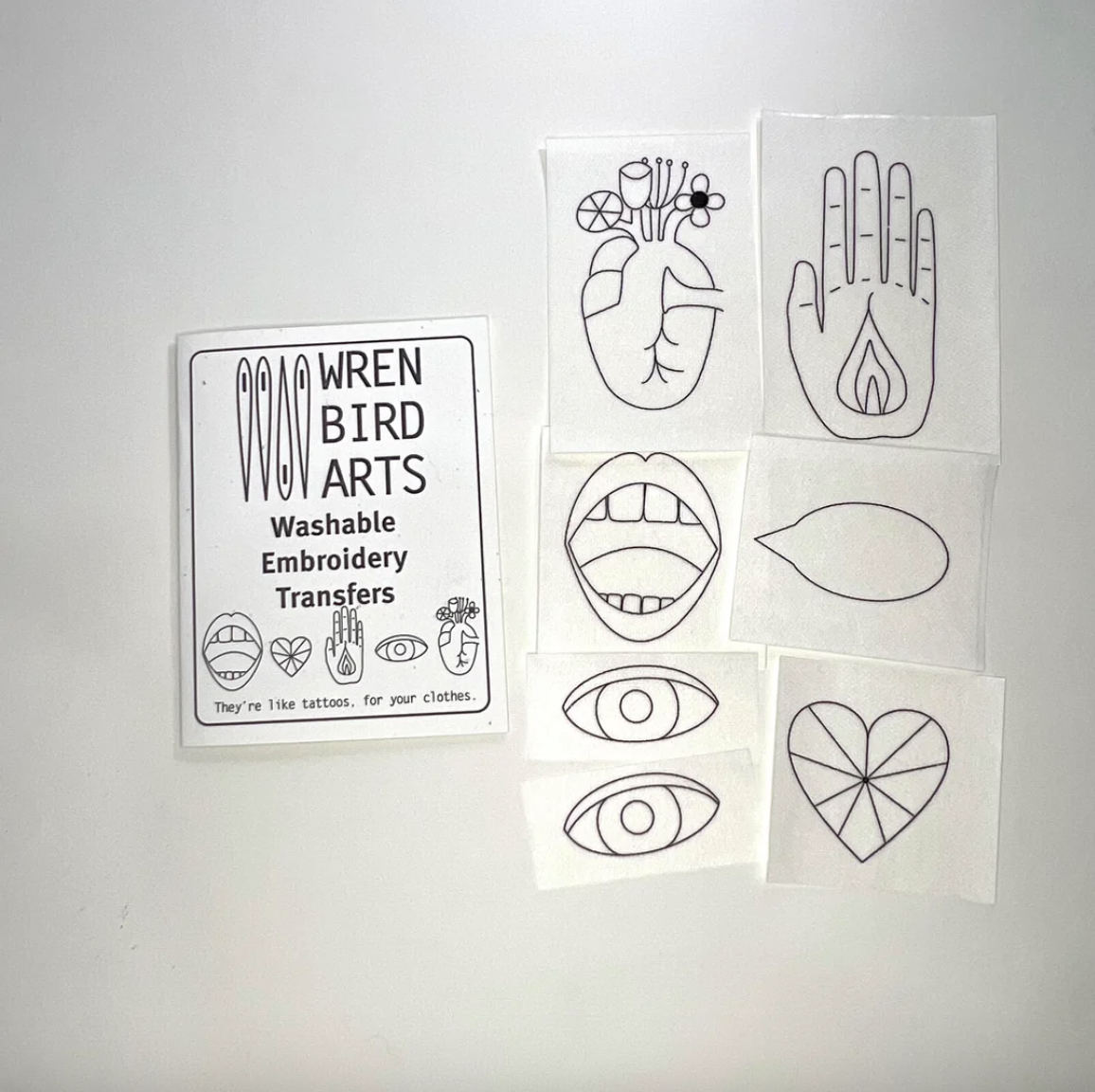 A set of white washable embroidery transfer sheets from Wren Bird Arts, featuring black line-art designs including a heart, hand, lips, eyes, and a heart motif, displayed next to a product label on a light gray background.