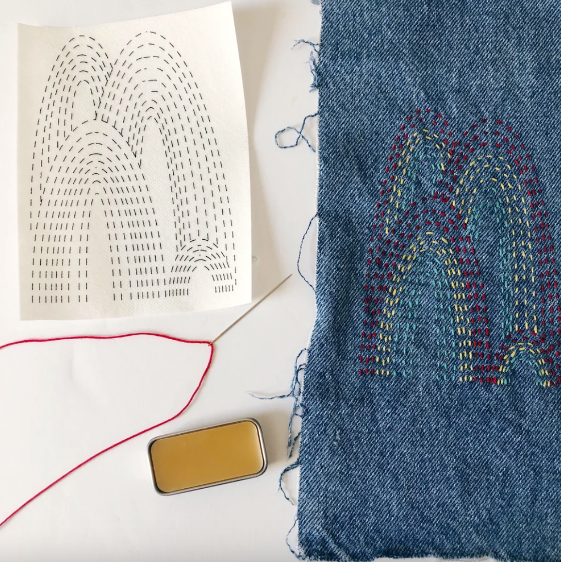 A stitched up example of a mending transfer on denim with a needle, thread, and tin of thread gloss. 