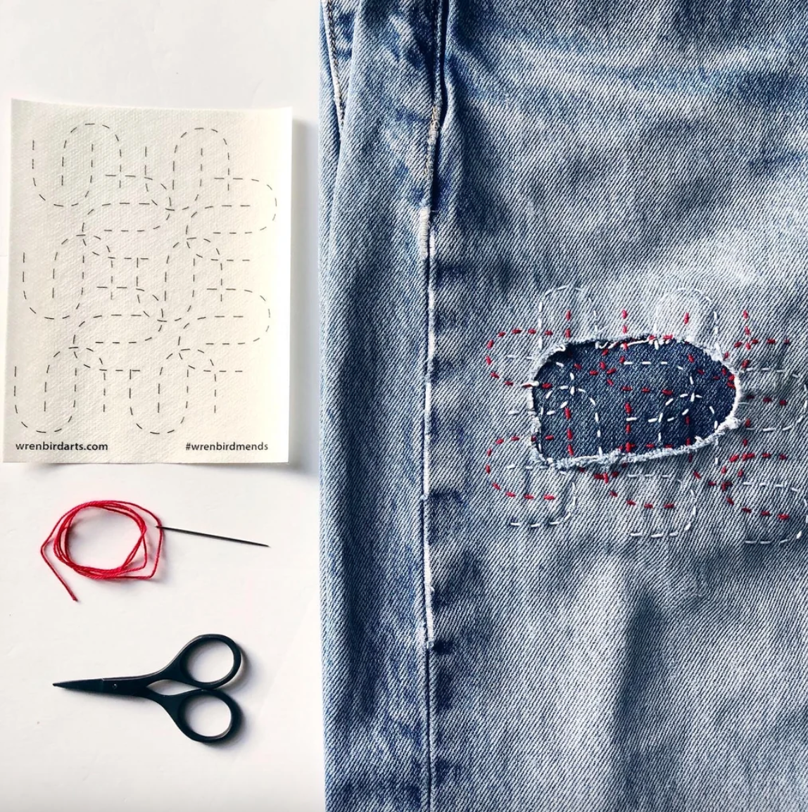 A stitched up example of one of the mending patterns on a pair of jeans with a needle and thread and a pair of scissors. 