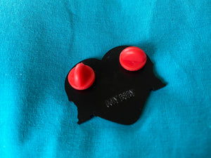 The back of the enamel pin. The pin is laying on a blue fabric background. It has two red coordinating rubber stoppers. The brand "Ugly Baby" is stamped on the back. 