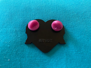 The back of the enamel pin. The pin is laying on a blue fabric background. It has two hot pink coordinating rubber stoppers. The brand "Ugly Baby" is stamped on the back. 