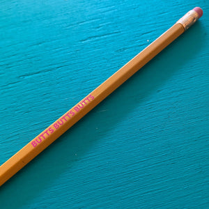A yellow pencil with a pink eraser is laying on a wood background painted turquoise. The pencil says "BUTTS BUTTS BUTTS" in bright pink text. 