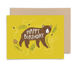 A mustard yellow card shows an image of a brown bear with rosy cheeks. It says "Happy Birthday" across his body. Bees and a hive are in the background along with some greenery. The card is shown on a white background with a Kraft colored envelope behind it.