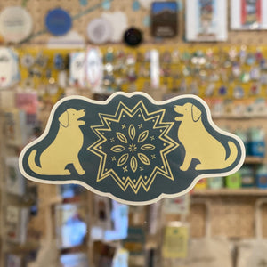 A folk art style sticker with two lab dogs sitting facing each other. A folk art design is between then. The sticker is blue and off-white. 