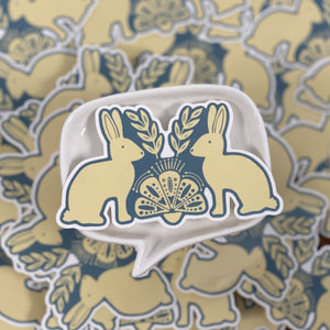 A blue and off-white die cut vinyl sticker by Ugly Baby. It is a folk art design of two rabbits facing each other. There is a folk art design between them.