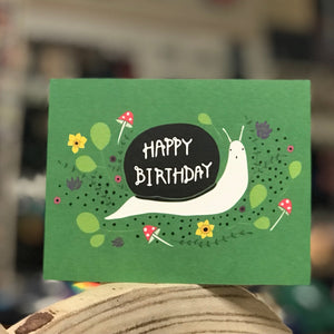 A greeting card with a bright green background. A surprised looking snail is carrying a shell that says "Happy Birthday" across his back. There are tiny mushrooms and illustrated flowers and leaves in the background. The card is sitting atop a piece of wood and the background of the photo is blurry. 
