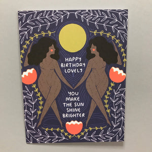 Two women with dark skin and long dark hair are holding hands. There is an intricate floral pattern drawn in the background surrounding a bright, yellow sun. The text says, "Happy Birthday Lovely. You Make the Sun Shine Brighter."
