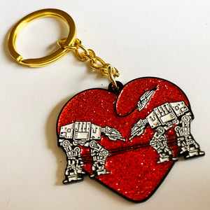 A glittery red keychain with gold hardware featuring a design of two robots surrounded by a heart. 