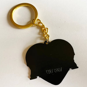 The back of a heart-shaped keychain with gold hardware. It is black and has the text "ugly baby" stamped into it. 