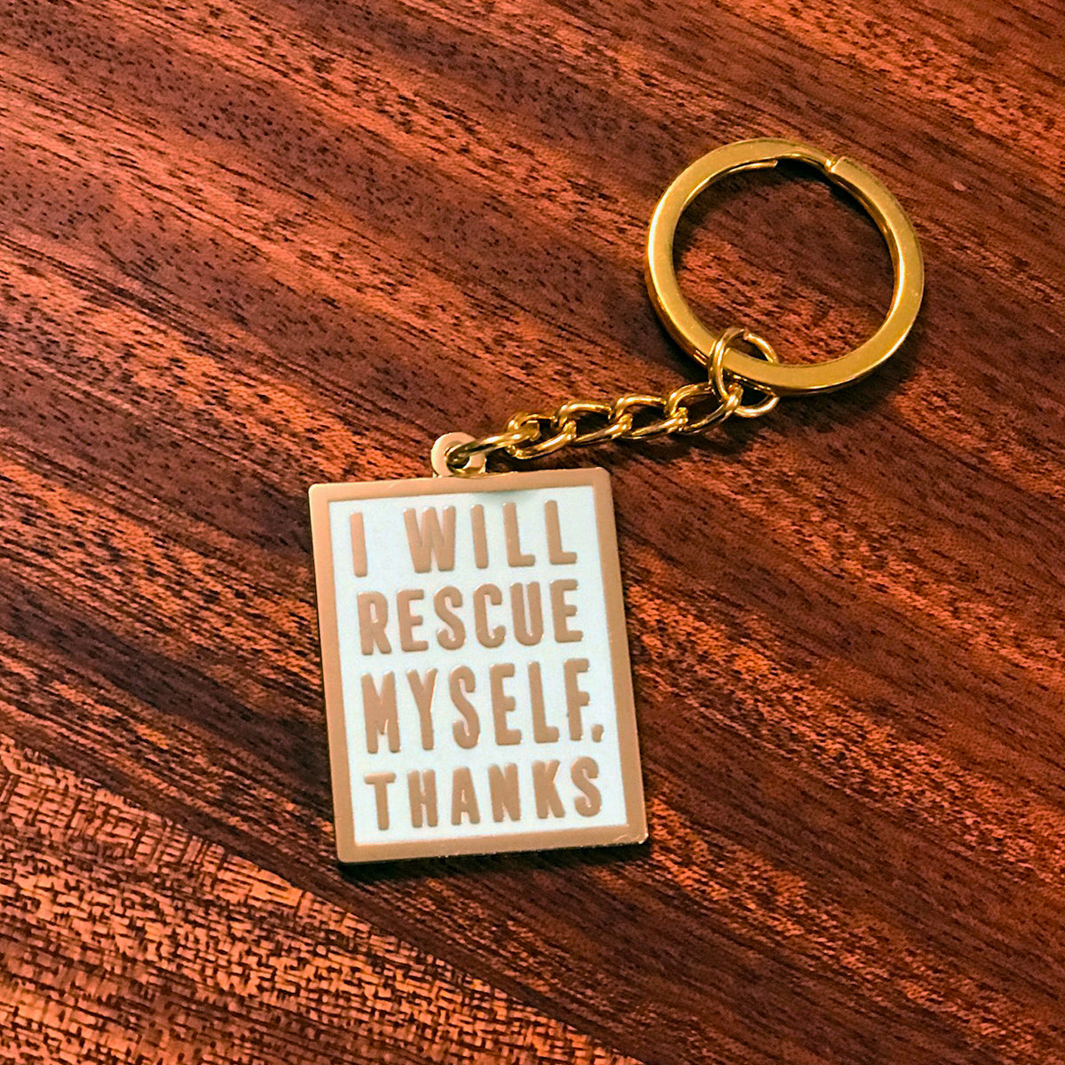A mint colored keychain with gold hardware and gold text that says "I will rescue myself, thanks" against a wooden background. 