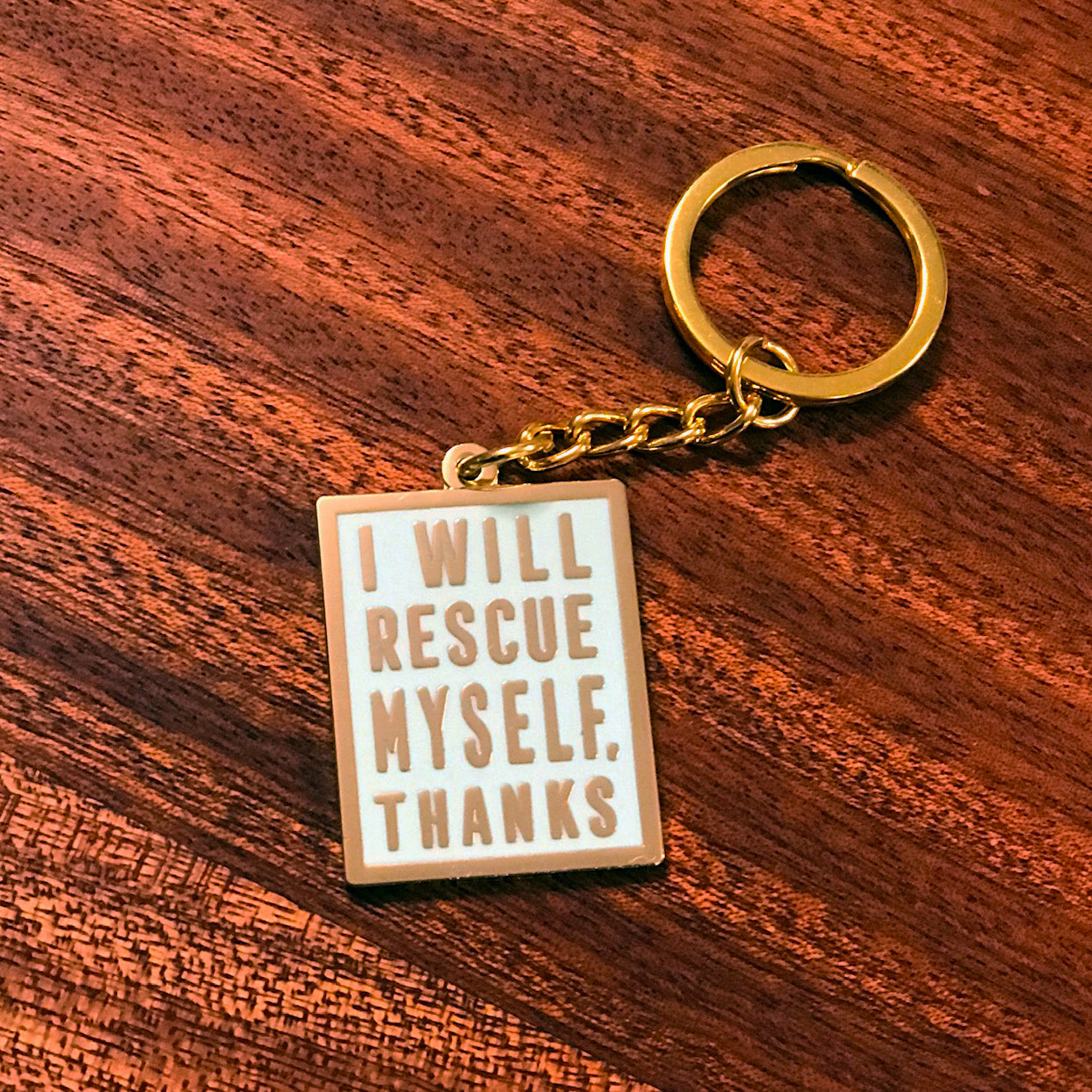 A mint colored keychain with gold hardware and gold text that says "I will rescue myself, thanks" against a wooden background. 