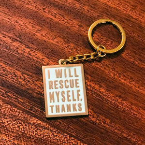 A mint colored keychain with gold hardware and gold text that says "I will rescue myself, thanks" against a wooden background. 